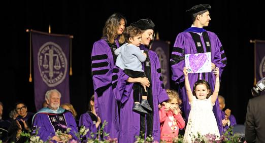Graduate accepting their diploma with their children, one of whom is holding a hand-made drawing of a purple heart